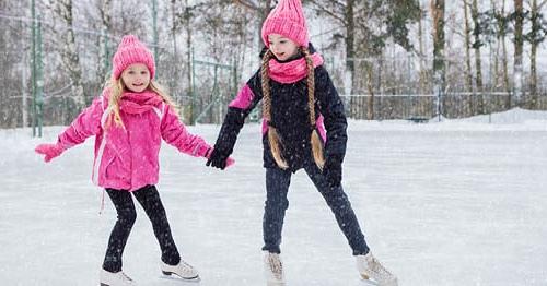 Two young females on an ice rink in pink coats ice skating and enjoying a snowy winter day. 